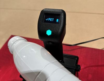 Kleiner schwarzer Kunststoffscanner. Auf dem Scanner liegt eine weiße Plastikflasche. Der Scanner identifiziert die Flasche als PET auf dem Display