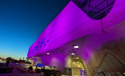 Nachtaufnahme vom phaeno Gebäude von außen, violett beleuchtet, im Hintergrund die Schornsteine von Volkswagen
